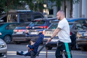 Фото мужчины с татуировкой на предплечье, толкающего детскую коляску по улице. Видна часть рисунка татуировки на открытой коже.
