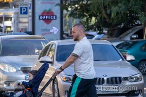 Фото мужчины с татуировкой на предплечье, толкающего детскую коляску по улице. Видна часть рисунка татуировки на открытой коже.