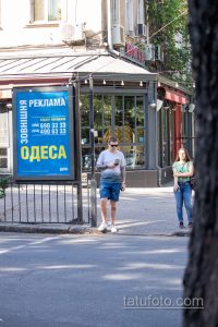 Фото тату на ноге мужчины, стоящего на улице. Видна татуировка на икре, повседневная одежда, городская обстановка.