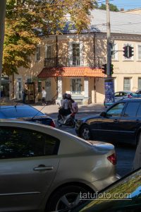 Фотография мотоциклиста с татуировкой на плече, едущего по улице. Видна часть рисунка татуировки, выполненной в черном цвете.