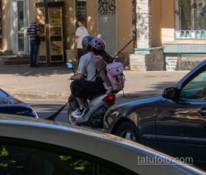 Фотография мотоциклиста с татуировкой на плече, едущего по улице. Видна часть рисунка татуировки, выполненной в черном цвете.