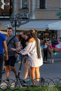 Фотография женщины с татуировкой на плече, едущей на электросамокате по городской улице. Видна часть рисунка татуировки, выполненной в черном цвете.