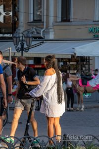 Фотография женщины с татуировкой на плече, едущей на электросамокате по городской улице. Видна часть рисунка татуировки, выполненной в черном цвете.