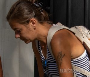 Фотография небольшой татуировки на плече женщины. Татуировка изображает стилизованный цветок или растительный орнамент.
