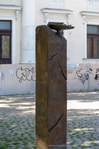 Фотография уличной татуировки на деревянном столбе, покрытом надписями и узорами. Изображение сделано на улице при дневном свете.