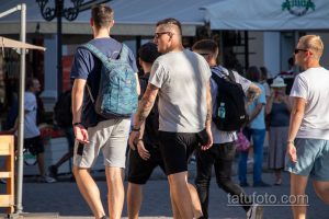 Фото мужчины с татуировками на руках и ногах, идущего в толпе людей. Татуировки в стиле олдскул с черепами, розами и другими элементами.
