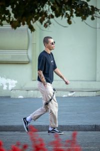 Фотография молодого человека в черной футболке и бежевых брюках с татуировкой на предплечье, идущего по улице.