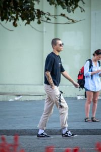 Фотография молодого человека в черной футболке и бежевых брюках с татуировкой на предплечье, идущего по улице.
