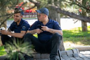 Фотография двух полицейских, сидящих под деревом и смотрящих в телефоны. На предплечьях видны татуировки с надписями.