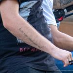 Фото тату на руке мужчины с надписью, выполненной черными чернилами. Надпись выглядит как цитата или личное послание. Уличная фотография.