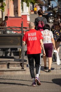 Фото тату на спине мужчины в красной футболке с надписью. Татуировка выполнена в черном цвете и представляет собой небольшую композицию. Уличная фотография.
