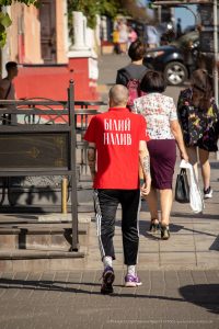 Фото тату на плече мужчины в красной футболке с надписью на спине. Татуировка выполнена в черном цвете и представляет собой небольшую композицию. Уличная фотография.