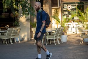 Фото мужчины с татуировками на руках и ногах, идущего по улице города. Видны детали рисунков татуировок и их стиль.