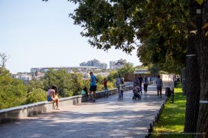 Фотография городской улицы с людьми, гуляющими по набережной. Вид на город и море на заднем плане. Яркий солнечный день.