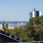 Фотография морского пейзажа с видом на город и отель с надписью 'Odessa'. Голубое небо, спокойное море и городская архитектура создают живописную картину.