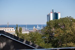 Фотография морского пейзажа с видом на город и отель с надписью 'Odessa'. Голубое небо, спокойное море и городская архитектура создают живописную картину.