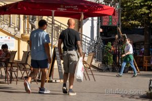 Фото мужчины с татуировкой на руке, стоящего на улице возле кафе. Видна часть татуировки, покрывающей руку.