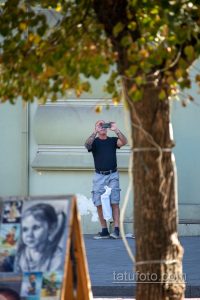 Фото мужчины с татуировкой на руке, фотографирующего на телефон. Видна детализированная татуировка на предплечье.