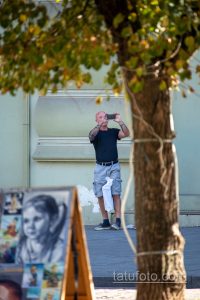 Фото мужчины с татуировкой на руке, фотографирующего на телефон. Видна детализированная татуировка на предплечье.