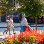 Фотография уличного художника в серебристой накидке и желтых брюках с украинским флагом, идущего по улице в Одессе, мимо прохожей.