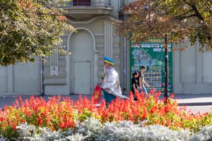 Фотография уличного художника в серебристой накидке и желтых брюках с украинской символикой, идущего по улице в окружении людей и цветов.