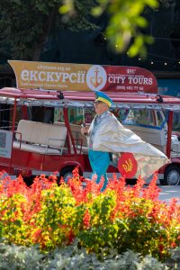 Фотография уличного художника с татуировкой на руке, стоящего рядом с красным туристическим автобусом в Одессе. Яркий костюм и динамичная поза привлекают внимание.