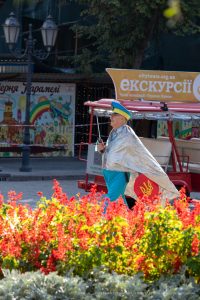 Фотография уличного художника в серебристой накидке и желтых брюках с украинским флагом, идущего по улице в Одессе.