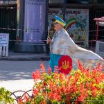 Фотография уличного художника в серебристой накидке и желтых брюках с украинским флагом, идущего по улице в Одессе.