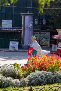Фотография уличного художника в серебристой накидке и желтых брюках с украинским флагом, идущего по улице в Одессе.