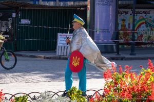 Фотография уличного художника в серебристой накидке и желтых брюках с украинским флагом, идущего по улице в Одессе.