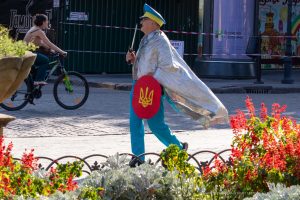 Фотография уличного художника в серебристой накидке и желтых брюках с украинским флагом, идущего по улице в Одессе, мимо велосипедистки.