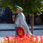 Фотография уличного художника в серебристой накидке и желтых брюках с украинским флагом, стоящего на улице в Одессе.