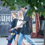 Фотография мужчины с татуировками на руке, идущего по улице с двумя девушками. Современный стиль одежды, солнечный день.
