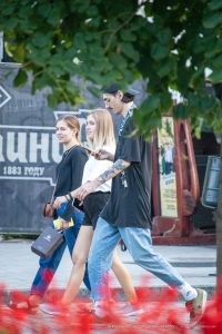 Фотография мужчины с татуировками на руке, идущего по улице с двумя девушками. Современный стиль одежды, солнечный день.