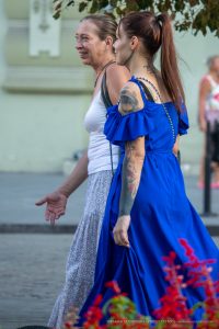 Фото тату на руке и шее женщины в синем платье, идущей рядом с другой женщиной. Детализированный рисунок татуировки, стиль стрит-арт.