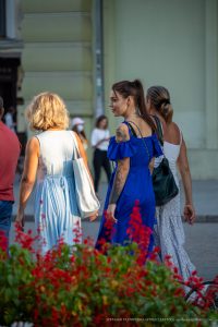 Фотография девушки с татуировками на плече и шее, идущей по улице в синем платье. Вид сзади, акцент на татуировках.