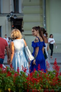 Фотография девушки с татуировками на плече и шее, идущей по улице в синем платье. Вид сзади, акцент на татуировках.