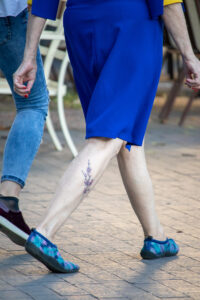 Фотография небольшой татуировки на лодыжке, изображающей веточку цветущей сакуры. Рисунок выполнен в нежных розовых и фиолетовых тонах с использованием тонких линий.