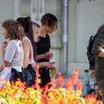 Фотография уличной татуировки: группа людей рассматривает работы мастера тату, портреты и эскизы на фоне ярких цветов и уличной обстановки.