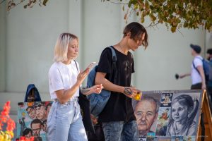 Фотография уличной татуировки: девушка и парень рассматривают работы мастера тату, портреты и эскизы на фоне ярких цветов.