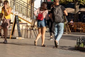 Фото тату на щиколотке девушки, идущей по улице. Небольшой рисунок татуировки в виде абстрактного символа, выполненный в черном цвете.