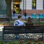 Фотография пары, сидящей на скамейке в парке. У парня на руке видна татуировка, а на футболке - принт с черепом.