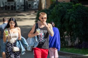 Фотография мужчины с татуировками на руках, одетого в черную майку с изображением черепа. Мужчина пьет напиток с трубочкой. Уличная фотография.
