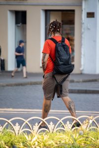 Фотография мужчины в оранжевой футболке с дредами и рюкзаком, на руках и ногах которого видны татуировки. Уличная татуировка, street tattoo.