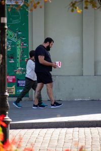 Фото тату на руке мужчины, идущего по улице. Видны татуировки на предплечье и кисти, выполненные в темных тонах. Уличная фотография.