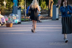 Фото тату на щиколотке девушки, идущей по улице. Небольшой рисунок татуировки в виде абстрактного символа, выполненный в черном цвете.