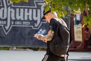 Фото тату на руке мужчины, запечатленного в момент курения. Рука покрыта детализированным рисунком татуировки в стиле олдскул. Мужчина одет в черную кепку и кожаную куртку.