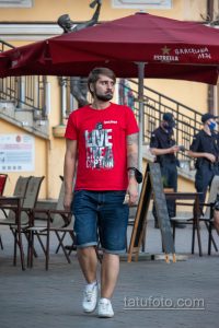 Фотография мужчины, идущего по улице в красной футболке. На его предплечье видна татуировка в черно-серых тонах, изображающая сложный узор.
