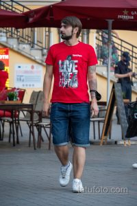 Фотография мужчины, идущего по улице в красной футболке. На его предплечье видна татуировка в черно-серых тонах, изображающая сложный узор.