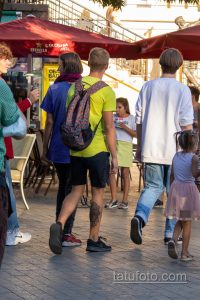 Фото татуировки дракона на голени мужчины, идущего по улице. Виден фрагмент тату, сочетающийся с повседневным стилем одежды.